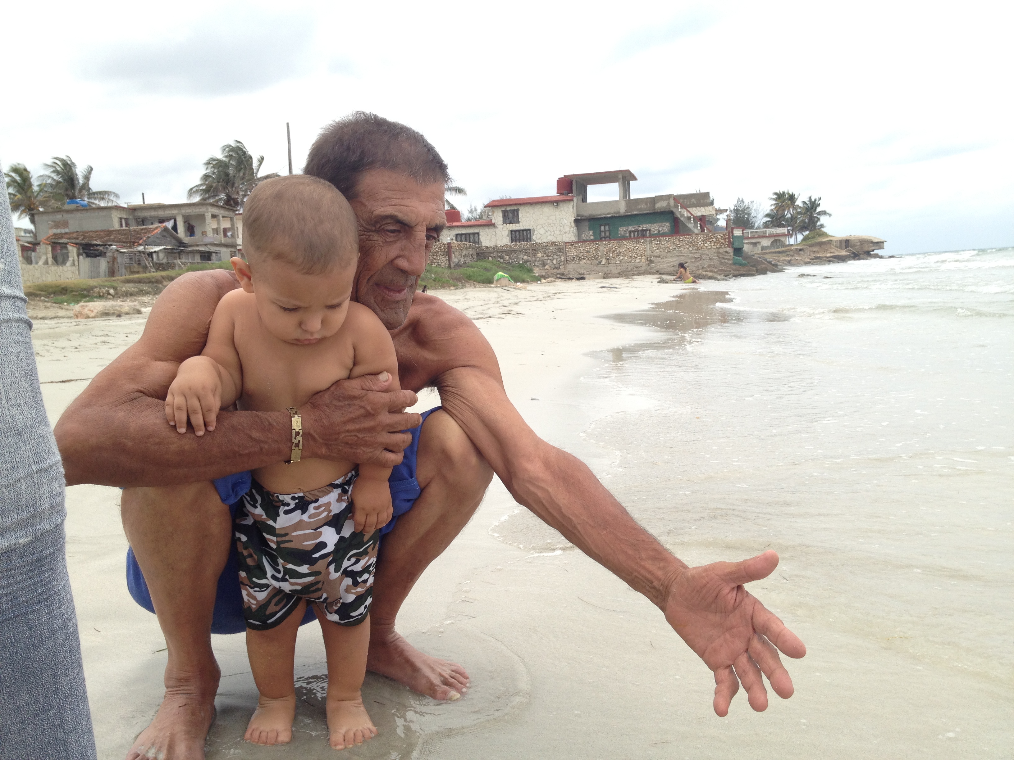 Papi agachado junto a un bebé en la playa, con las olas detrás.
