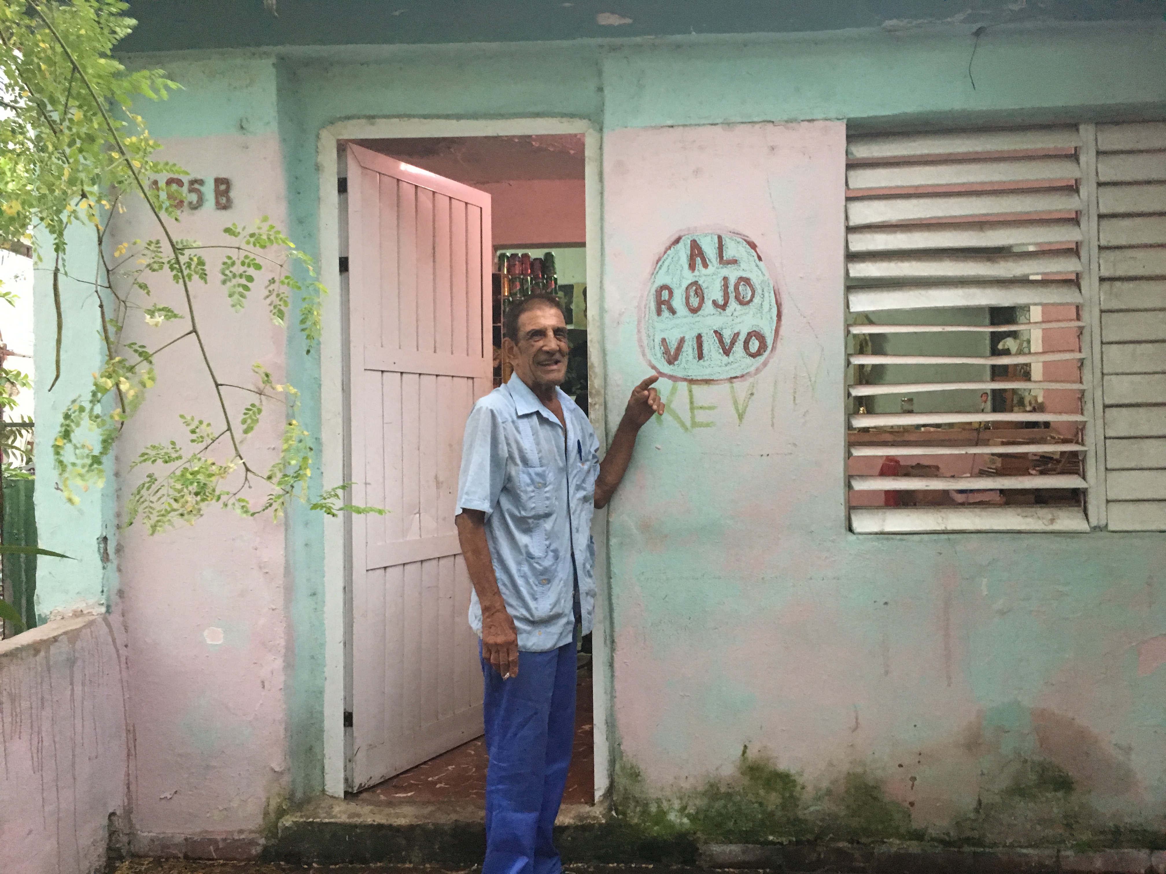 Papi standing in the doorway of his pink and mint-green house in Cuba, pointing at a hand-painted sign that reads 'AL ROJO VIVO' on the wall beside him.