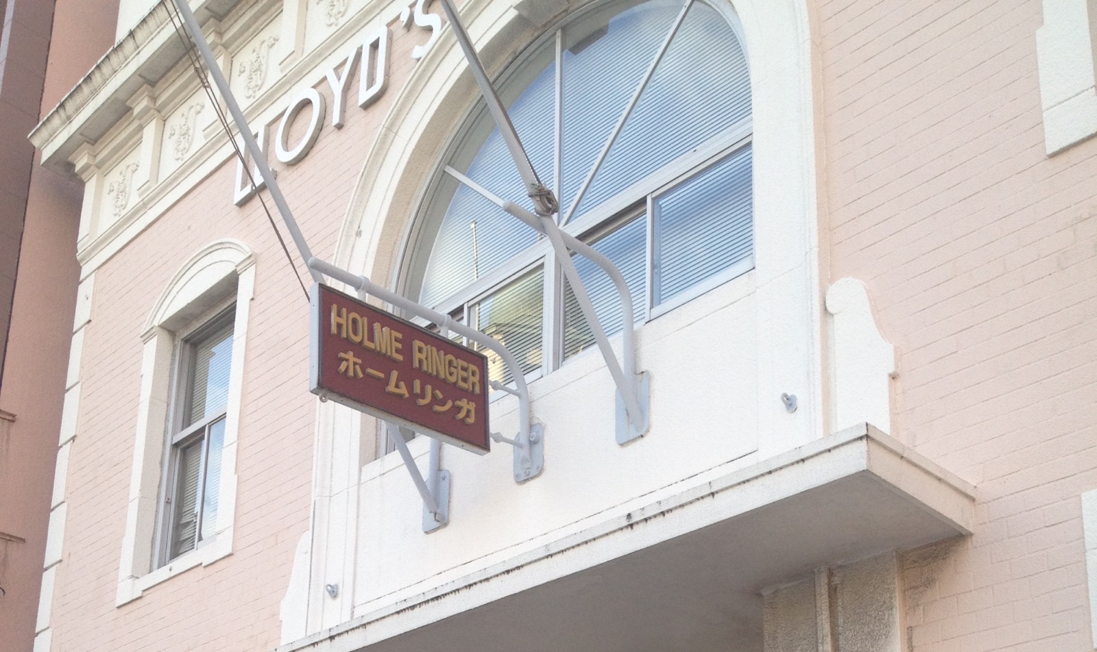 Un letrero colgante con las palabras HOLME RINGER (y el mismo nombre en katakana) frente al edificio de ladrillo rosado de Mojiko, con la palabra LLOYD'S en la fachada